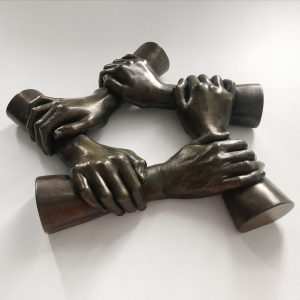 5 hands holding each other by the forearm in a hexagonal ring cast in bronze